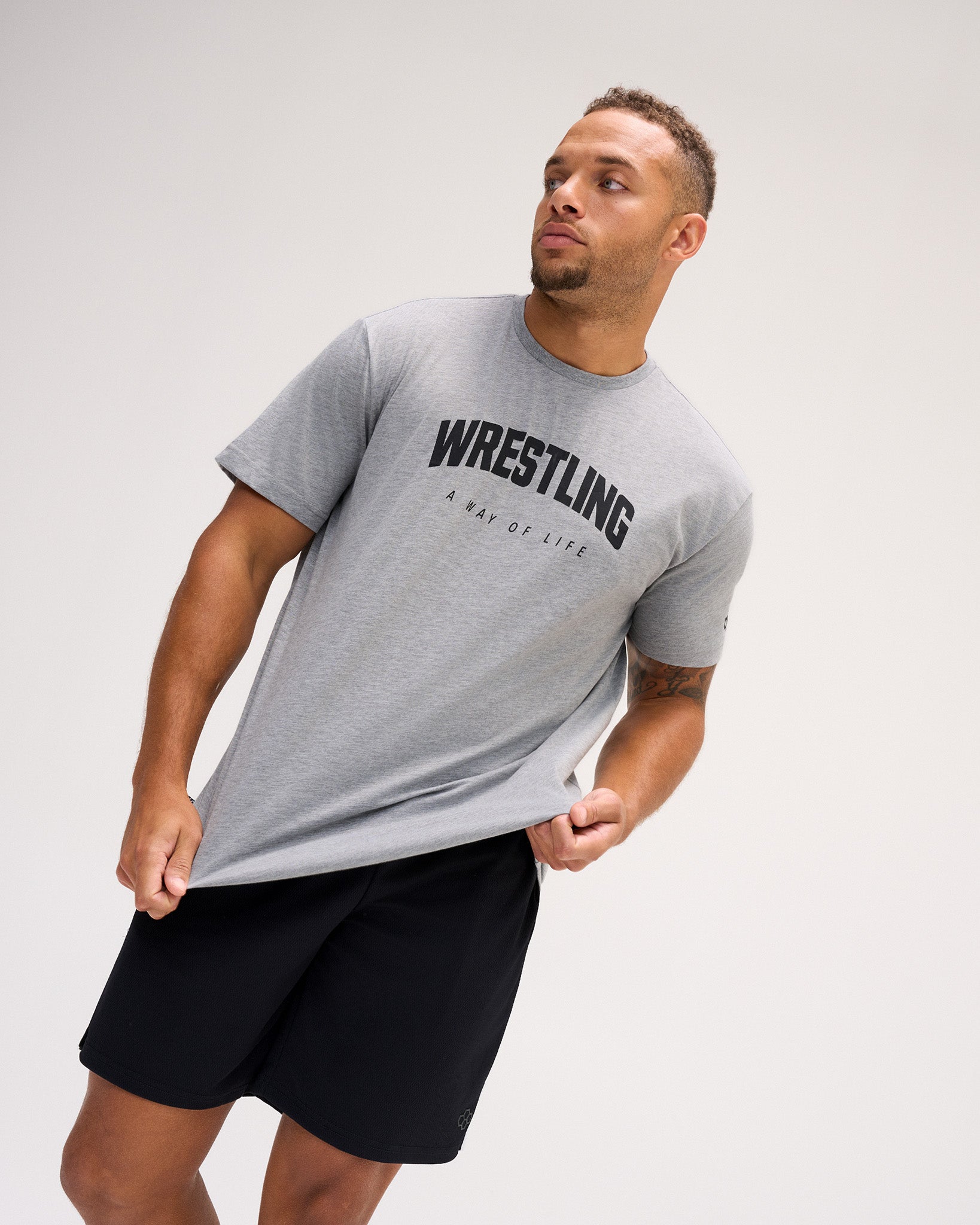 A man wearing a heather gray short-sleeve t-shirt with 'WRESTLING' printed in black across the chest, paired with black athletic shorts, against a light gray studio background.