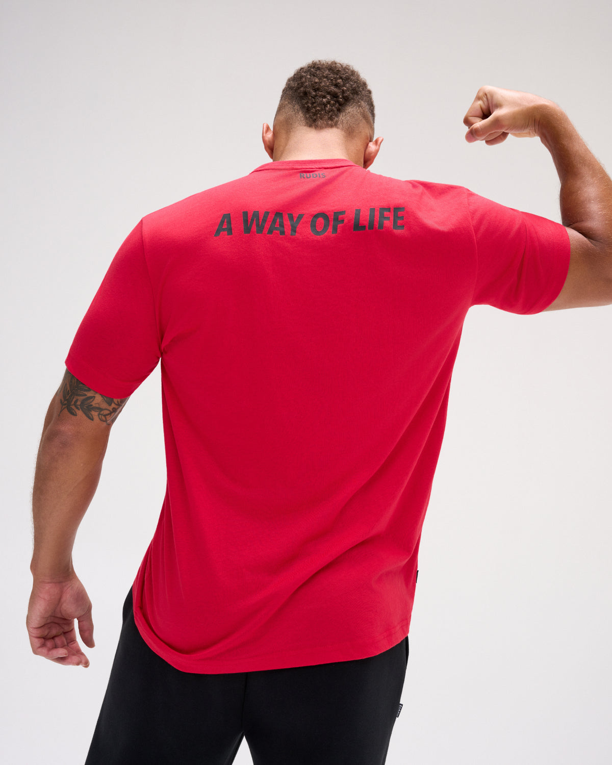 A man viewed from behind wearing a bright red athletic t-shirt with the text 'A WAY OF LIFE' printed in dark blue across the upper back, paired with black pants, against a light gray studio background.