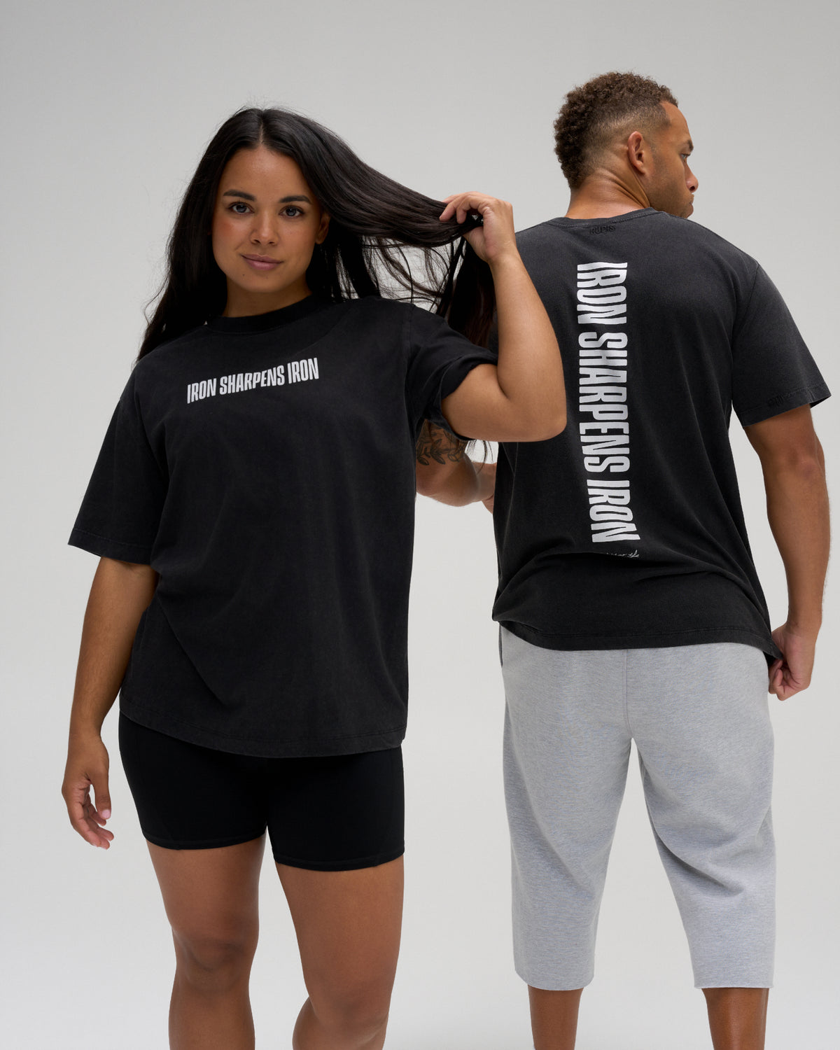 Two models wearing the RUDIS Iron Sharpens Iron Classic Cotton T-Shirt in Washed Black, showcasing the white front chest graphic, bold vertical back print and relaxed fit.