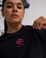 Close-up of a female model wearing the RUDIS Hex Classic Cotton T-Shirt in Black/Pink, highlighting the vibrant pink embroidered logo and black cotton fabric.