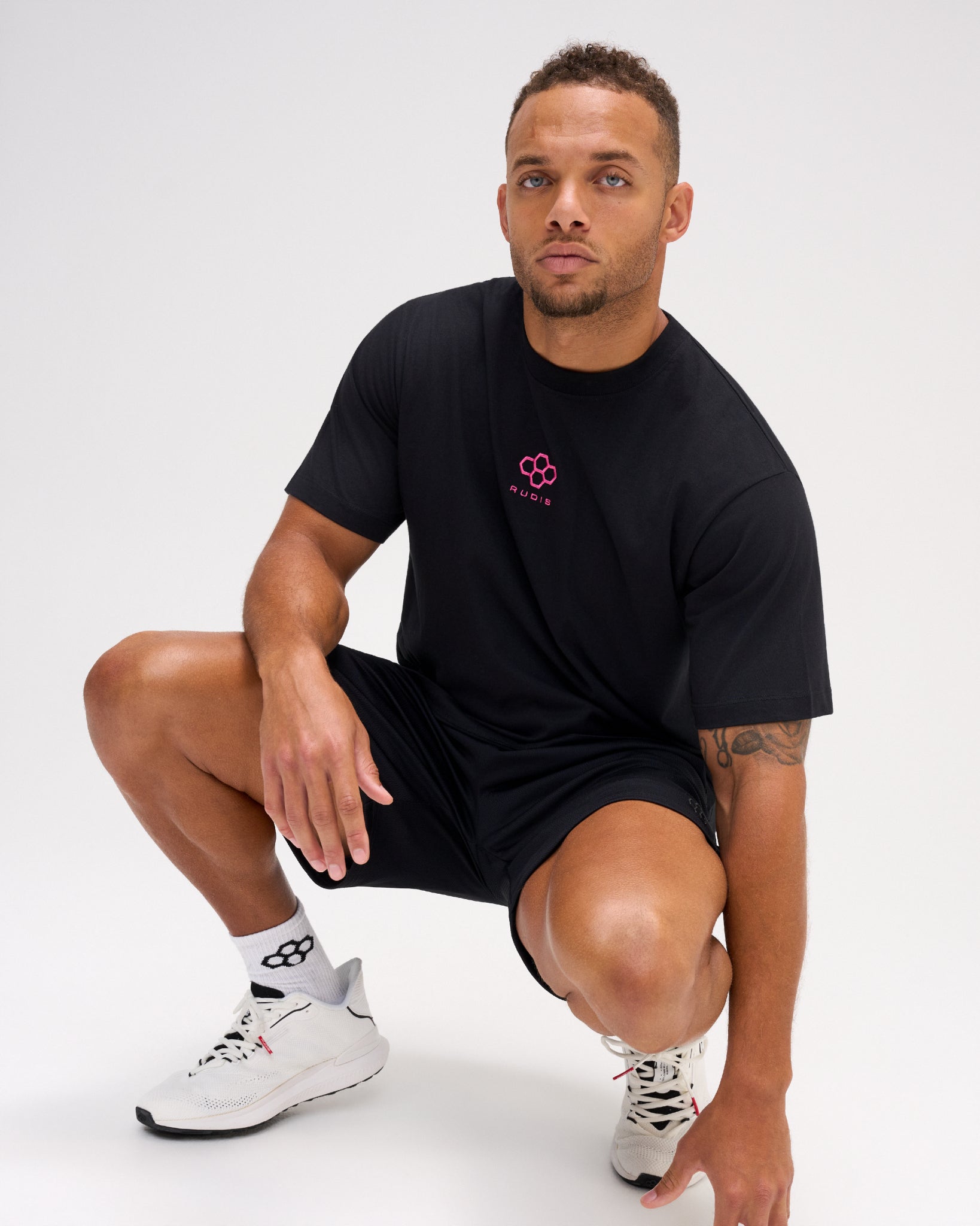 Male model crouching in the RUDIS Hex Classic Cotton T-Shirt in Black/Pink, showcasing the pink Hex chest embroidery and classic crewneck fit.