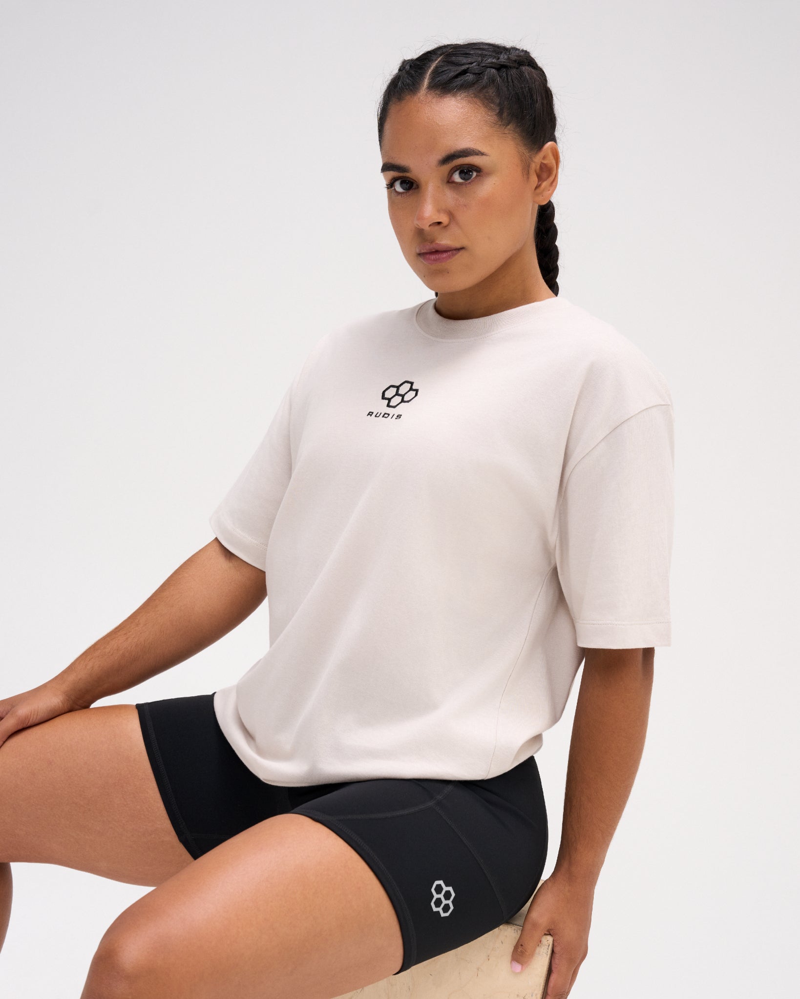 Female model seated in the RUDIS Hex Classic Cotton T-Shirt in Bone, highlighting the black embroidered Hex logo, crew neckline and relaxed fit.