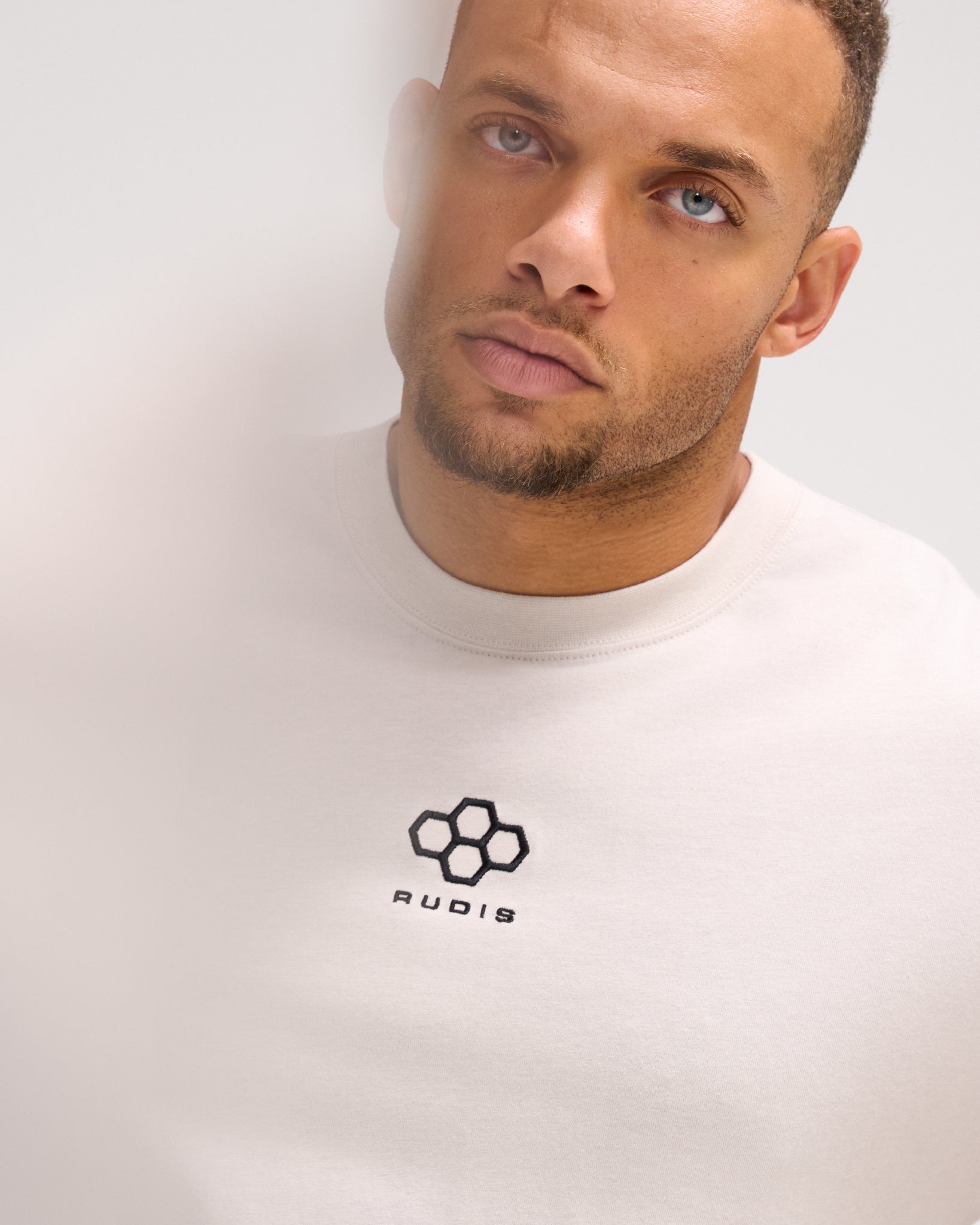 Close-up of a male model wearing the RUDIS Hex Classic Cotton T-Shirt in Bone, showcasing the black embroidered Hex logo and ribbed crew neckline.