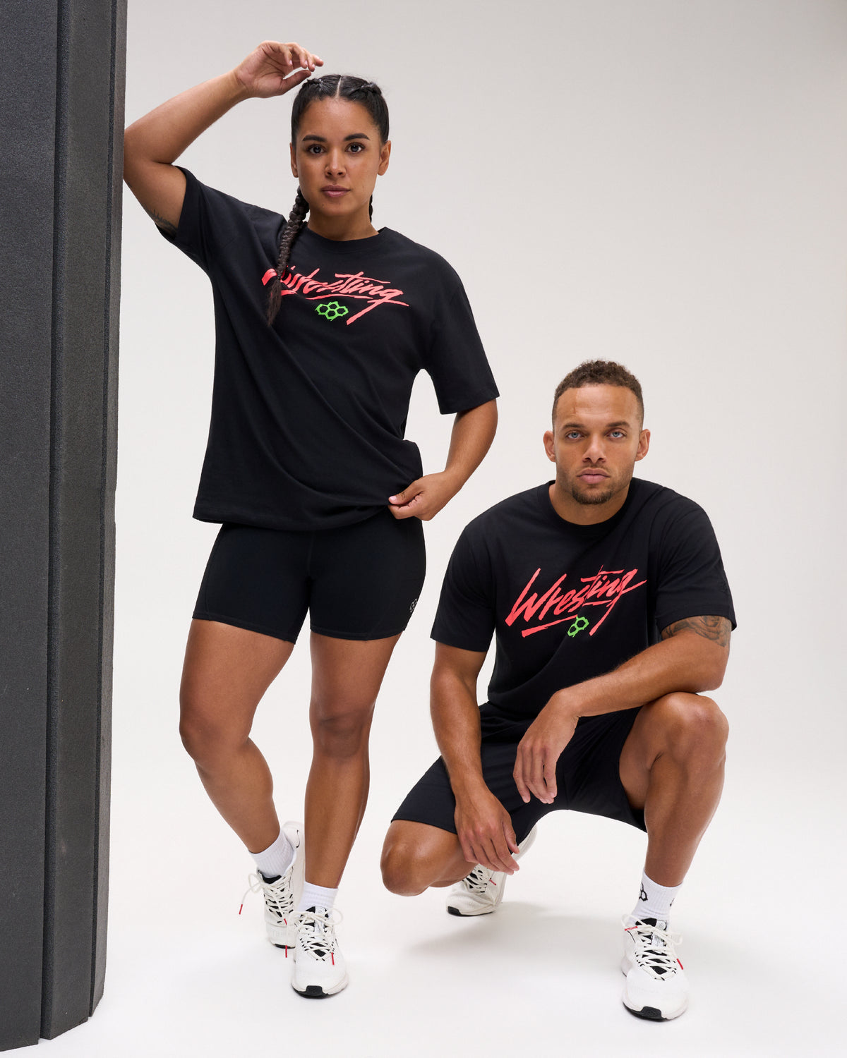Male and female models wearing the RUDIS Script Wrestling Classic Cotton T-Shirt in Black, featuring coral Wrestling script and a green Hex logo.
