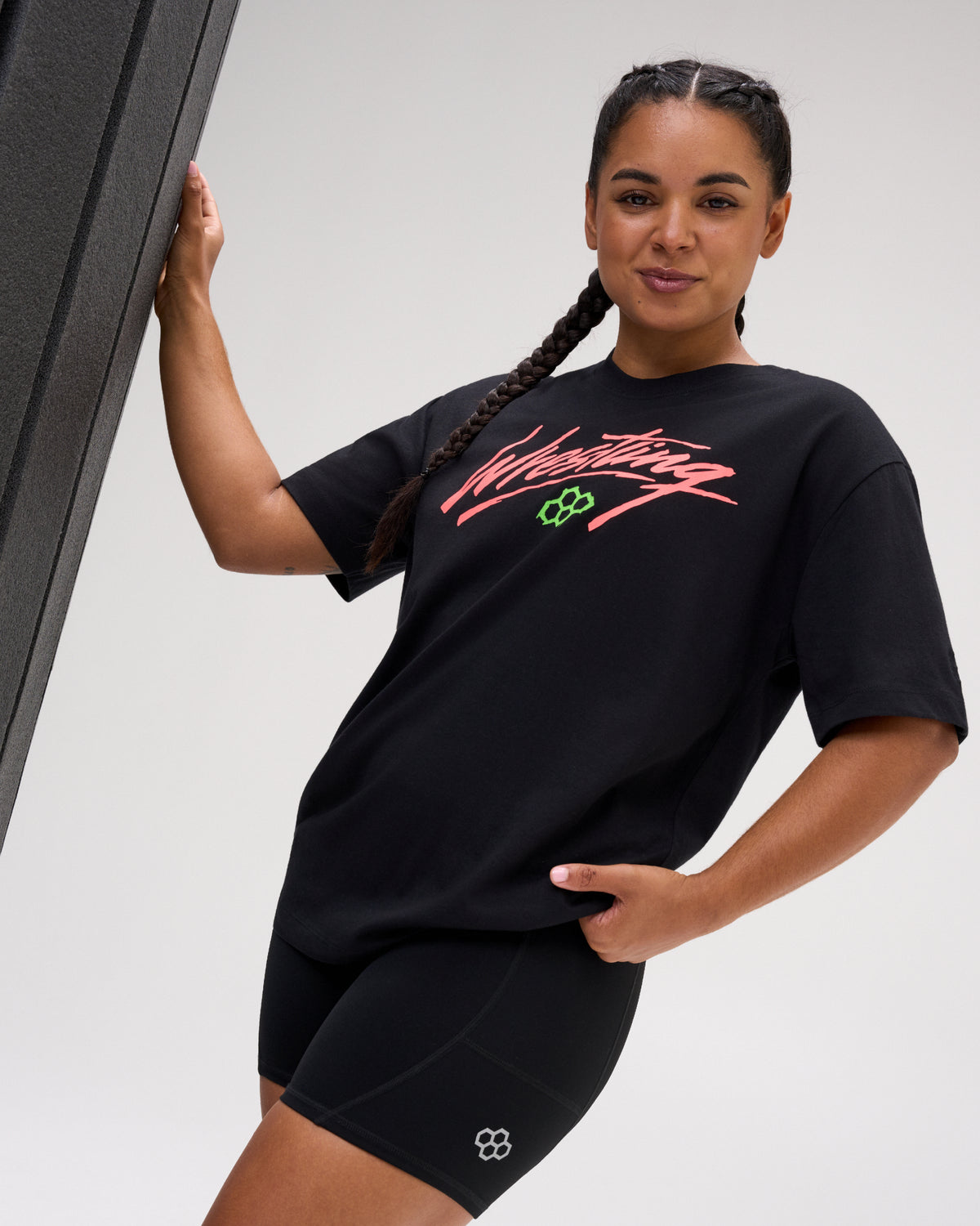 Female model wearing the RUDIS Script Wrestling Classic Cotton T-Shirt in Black with a bold coral Wrestling graphic, green Hex logo and relaxed fit.