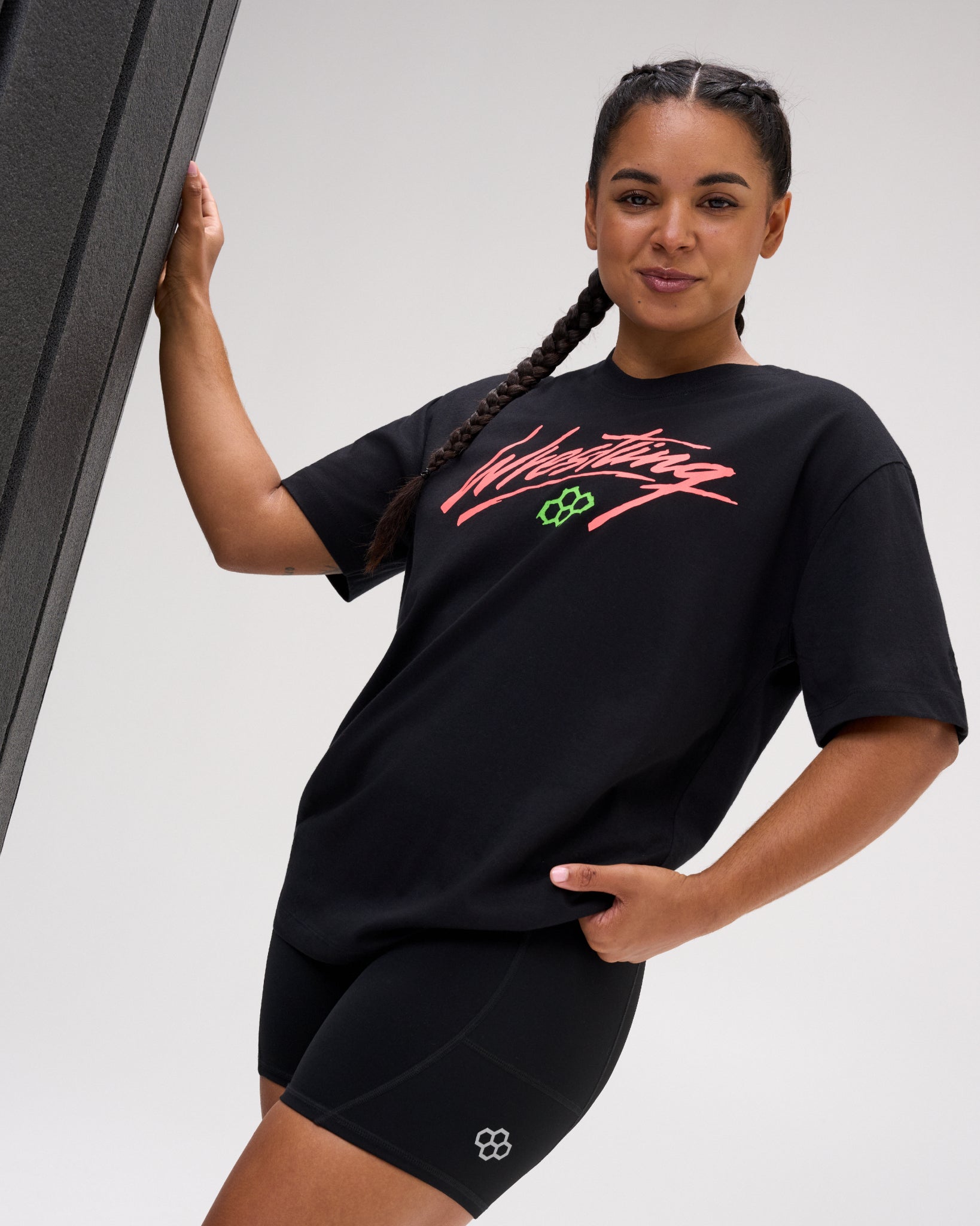 Female model wearing the RUDIS Script Wrestling Classic Cotton T-Shirt in Black with a bold coral Wrestling graphic, green Hex logo and relaxed fit.