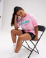 Seated female model wearing the RUDIS Script Wrestling Classic Cotton T-Shirt in Tie-Dye Pink, featuring teal Wrestling lettering and a pink Hex logo.