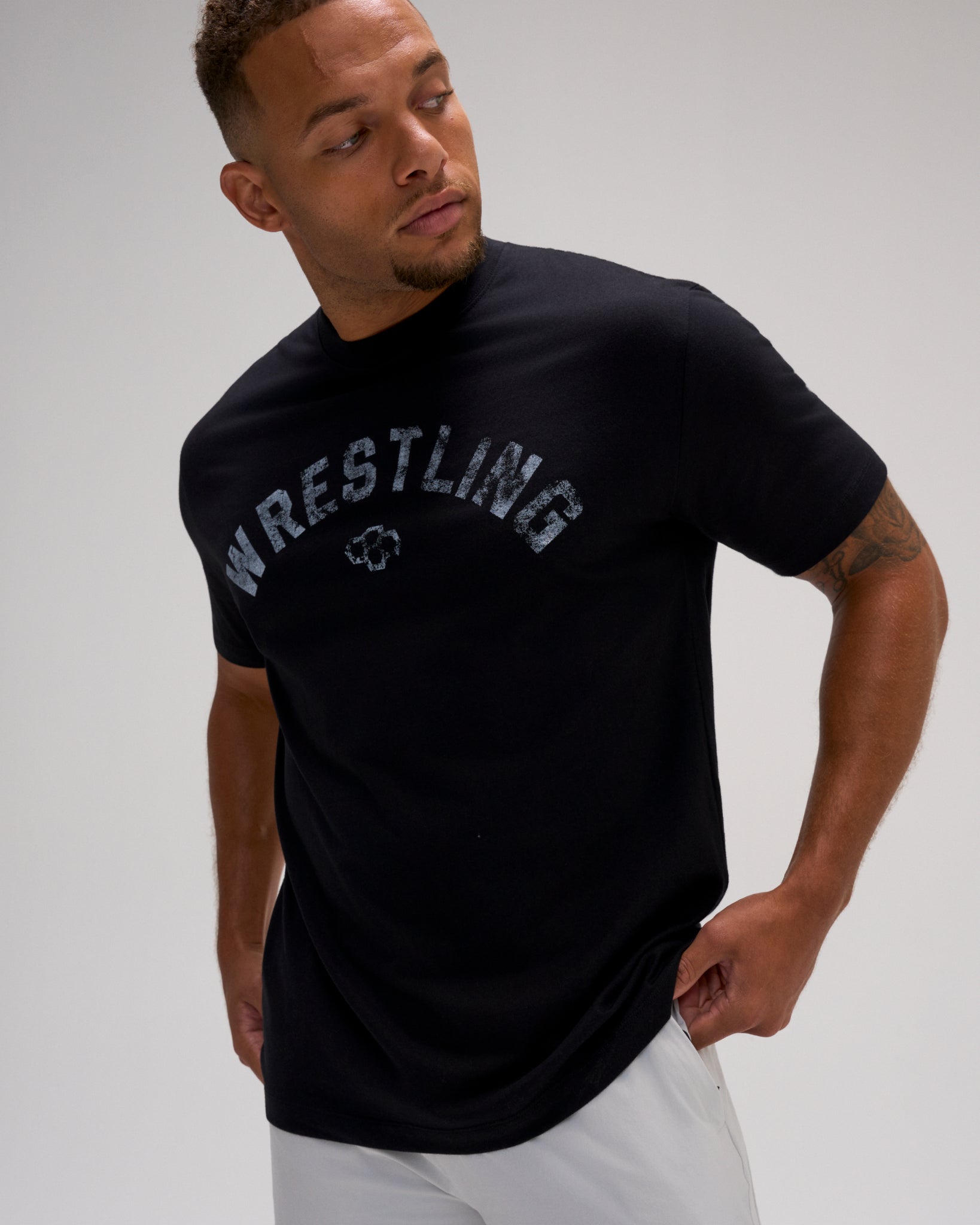 Male model wearing the RUDIS Wrestling Classic Cotton T-Shirt in Black with distressed gray arched Wrestling lettering and RUDIS Hex logo.