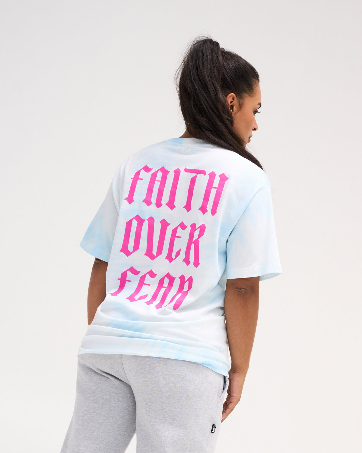 Female model wearing the RUDIS Faith Over Fear Classic Cotton T-Shirt in Blue Tie Dye, showing oversized pink gothic Faith Over Fear lettering on the back.