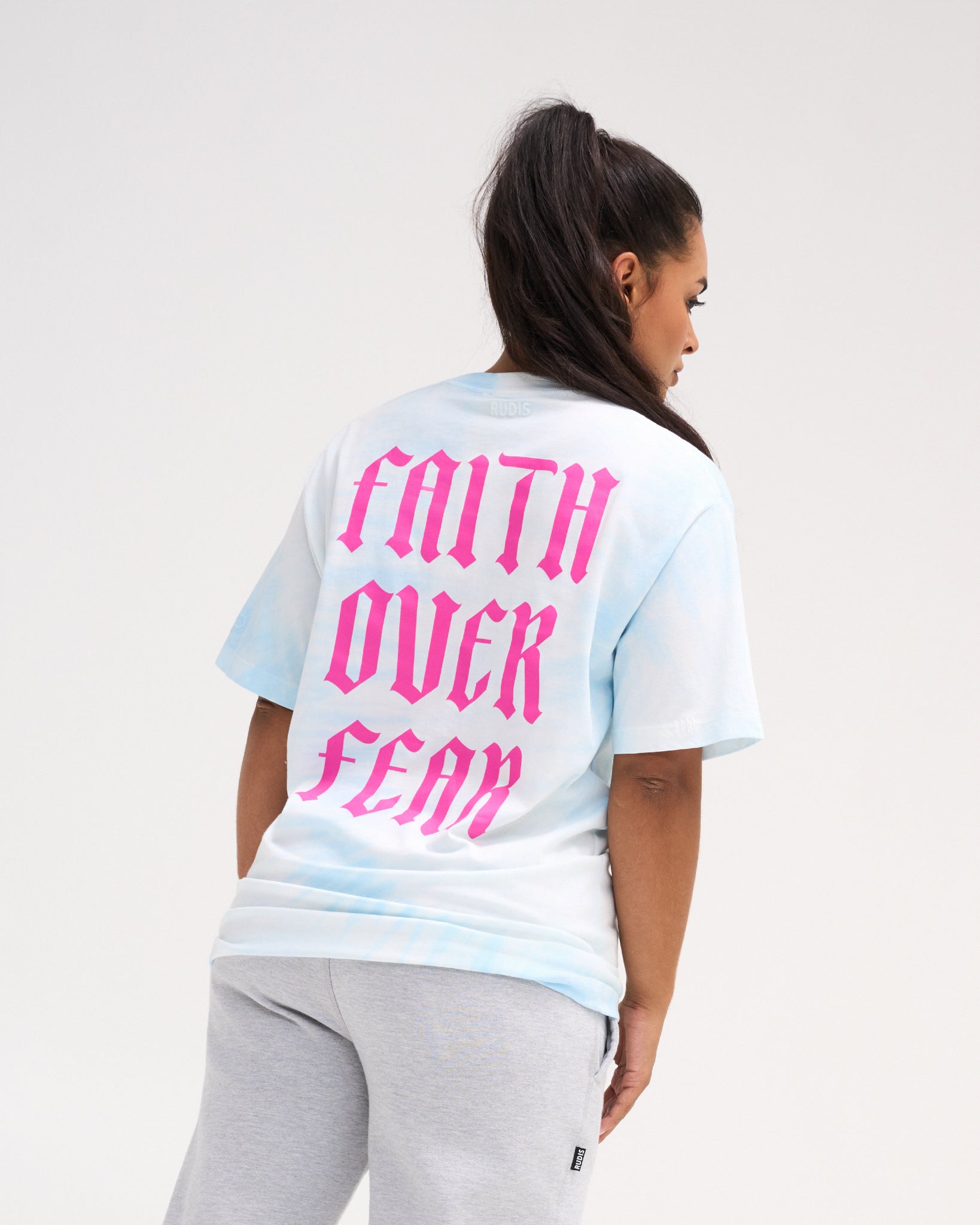Female model wearing the RUDIS Faith Over Fear Classic Cotton T-Shirt in Blue Tie Dye, showing oversized pink gothic Faith Over Fear lettering on the back.