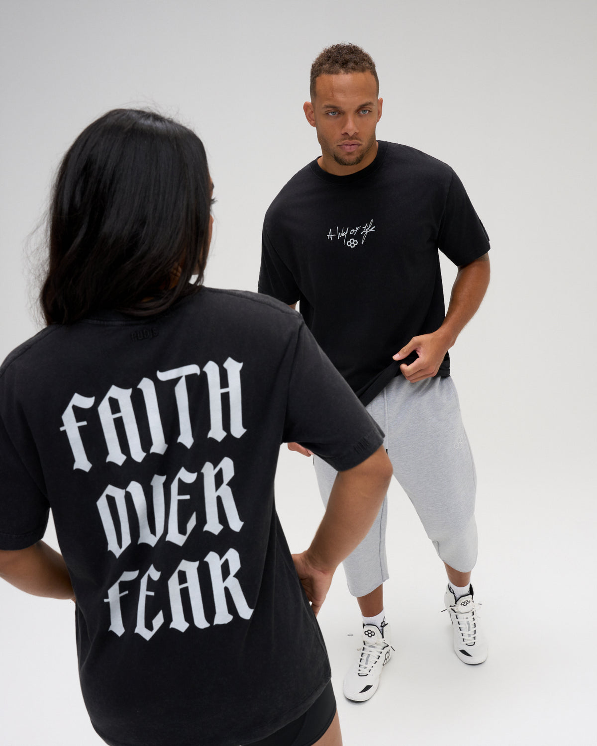 Male and female models wearing the RUDIS Faith Over Fear Classic Cotton T-Shirt in Washed Black, showing white chest graphics and bold back lettering.
