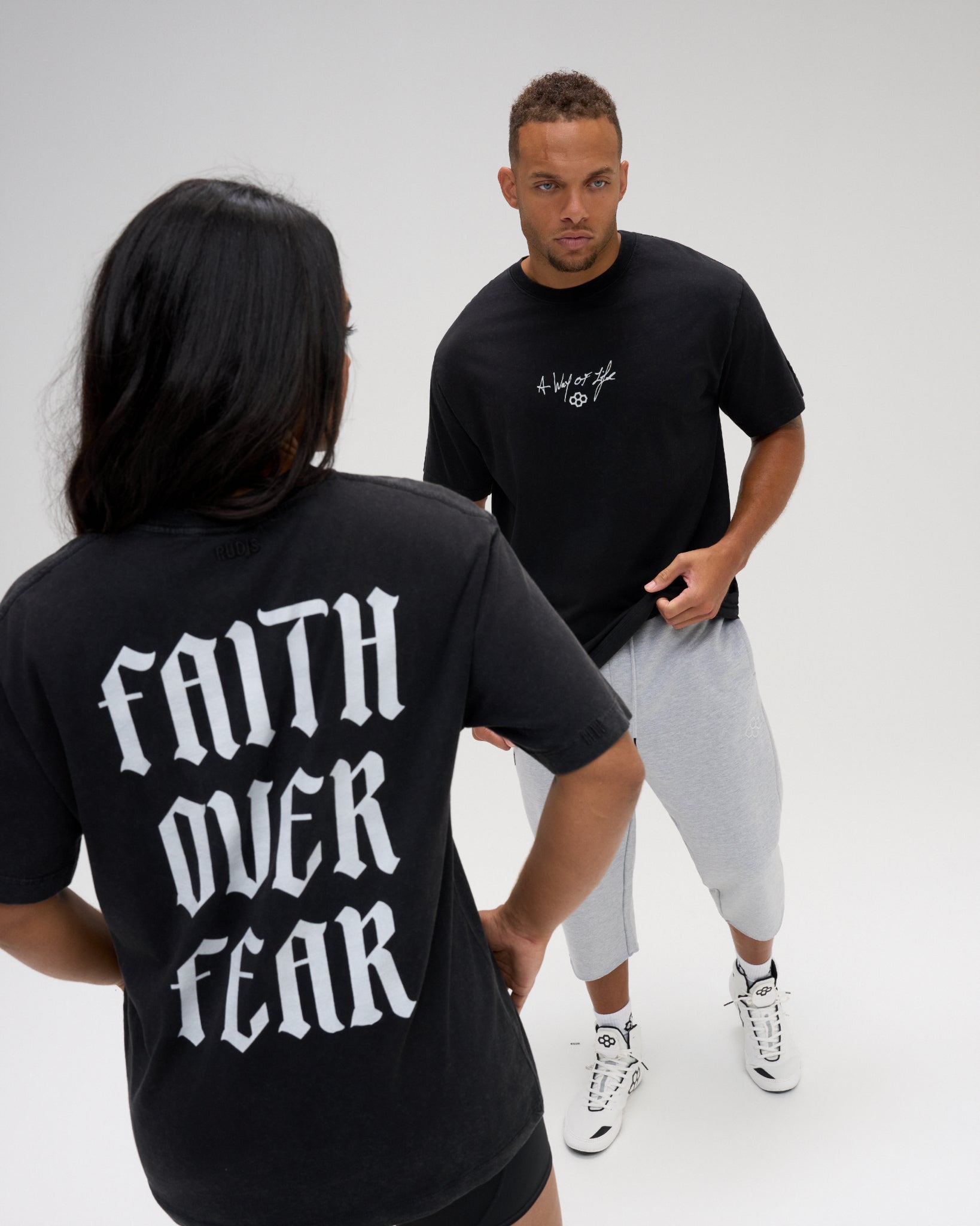 Male and female models wearing the RUDIS Faith Over Fear Classic Cotton T-Shirt in Washed Black, showing white chest graphics and bold back lettering.