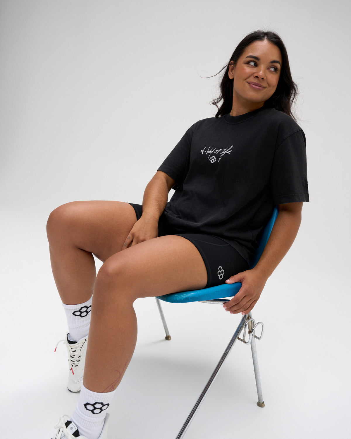 Female model seated in the RUDIS Faith Over Fear Classic Cotton T-Shirt in Washed Black, styled with black athletic shorts and a white chest graphic.