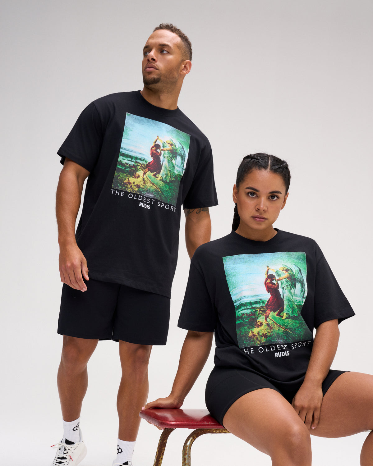 Male and female models wearing the RUDIS Divine Struggle Relaxed Cotton T-Shirt in Washed Black, featuring colorful wrestling-inspired artwork and The Oldest Sport graphic.