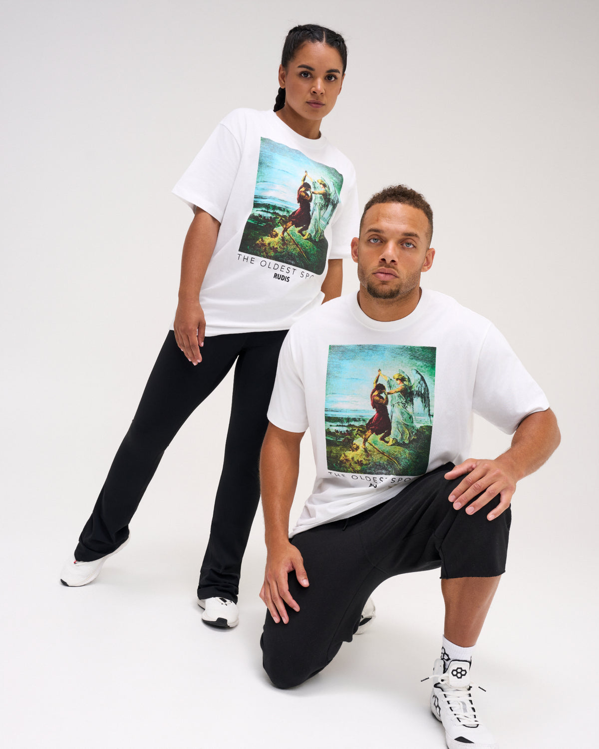 Male and female models wearing the RUDIS Divine Struggle Relaxed Cotton T-Shirt in White, featuring colorful artwork of two wrestling figures and The Oldest Sport graphic.
