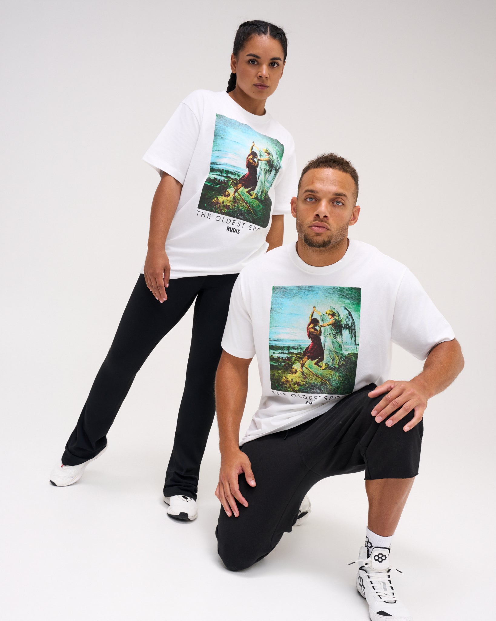 Male and female models wearing the RUDIS Divine Struggle Relaxed Cotton T-Shirt in White, featuring colorful artwork of two wrestling figures and The Oldest Sport graphic.