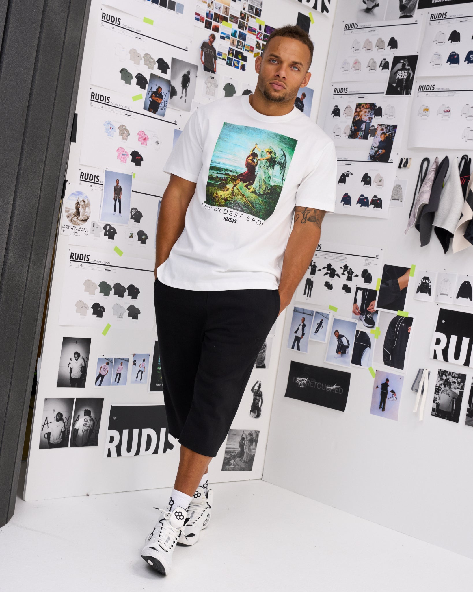 Male model wearing the RUDIS Divine Struggle Relaxed Cotton T-Shirt in White with vintage-style wrestling artwork and The Oldest Sport graphic, paired with black athletic shorts.