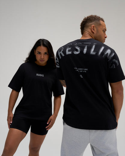 Male and female models wearing the RUDIS Wrestling Back Relaxed Cotton T-Shirt in Black, showing the minimal front RUDIS logo and oversized WRESTLING back graphic.