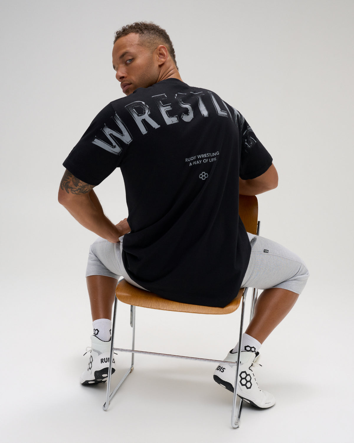 Male model seated wearing the RUDIS Wrestling Back Relaxed Cotton T-Shirt in Black, featuring a large distressed WRESTLING back print and A Way of Life message.