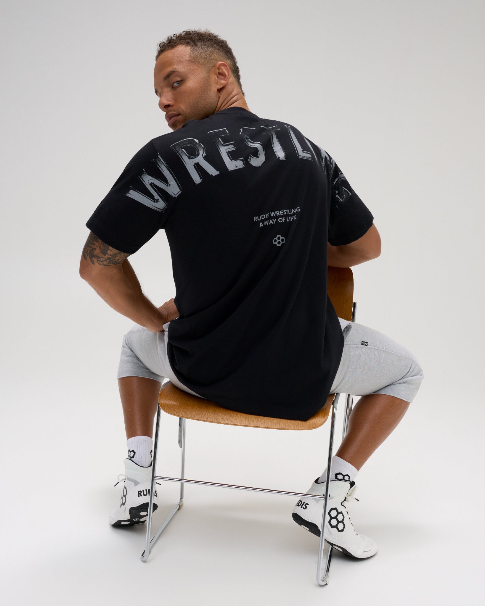 Male model seated wearing the RUDIS Wrestling Back Relaxed Cotton T-Shirt in Black, featuring a large distressed WRESTLING back print and A Way of Life message.