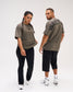 Male and female models wearing the RUDIS Wrestling Back Relaxed Cotton T-Shirt in Dark Olive, displaying the black RUDIS chest logo and bold WRESTLING back lettering.