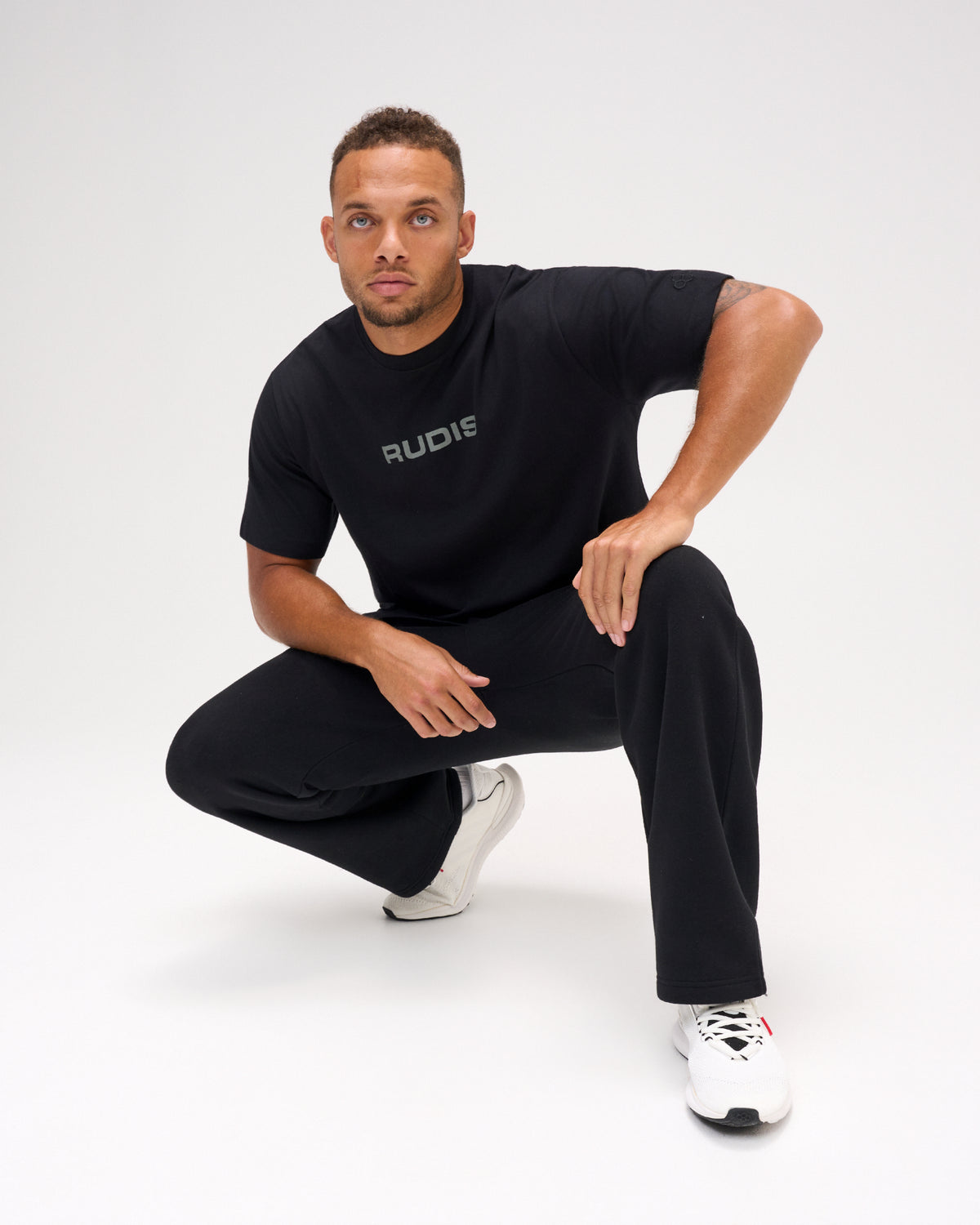 Male model crouching in a black RUDIS Relaxed Cotton T-Shirt featuring a gray RUDIS chest logo, paired with black pants and white shoes.