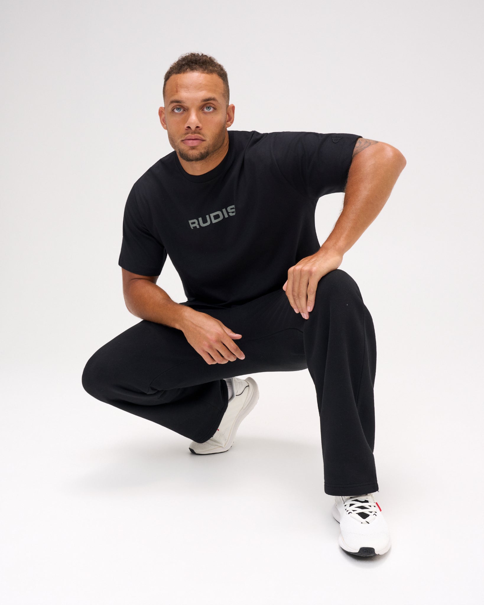 Male model crouching in a black RUDIS Relaxed Cotton T-Shirt featuring a gray RUDIS chest logo, paired with black pants and white shoes.