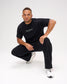 Male model crouching in a black RUDIS Relaxed Cotton T-Shirt featuring a gray RUDIS chest logo, paired with black pants and white shoes.