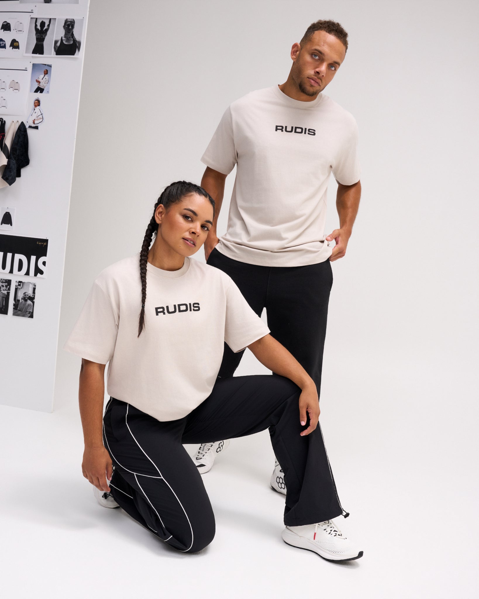 Male and female models wearing bone RUDIS Relaxed Cotton T-Shirts with black RUDIS chest logos, paired with black athletic pants.