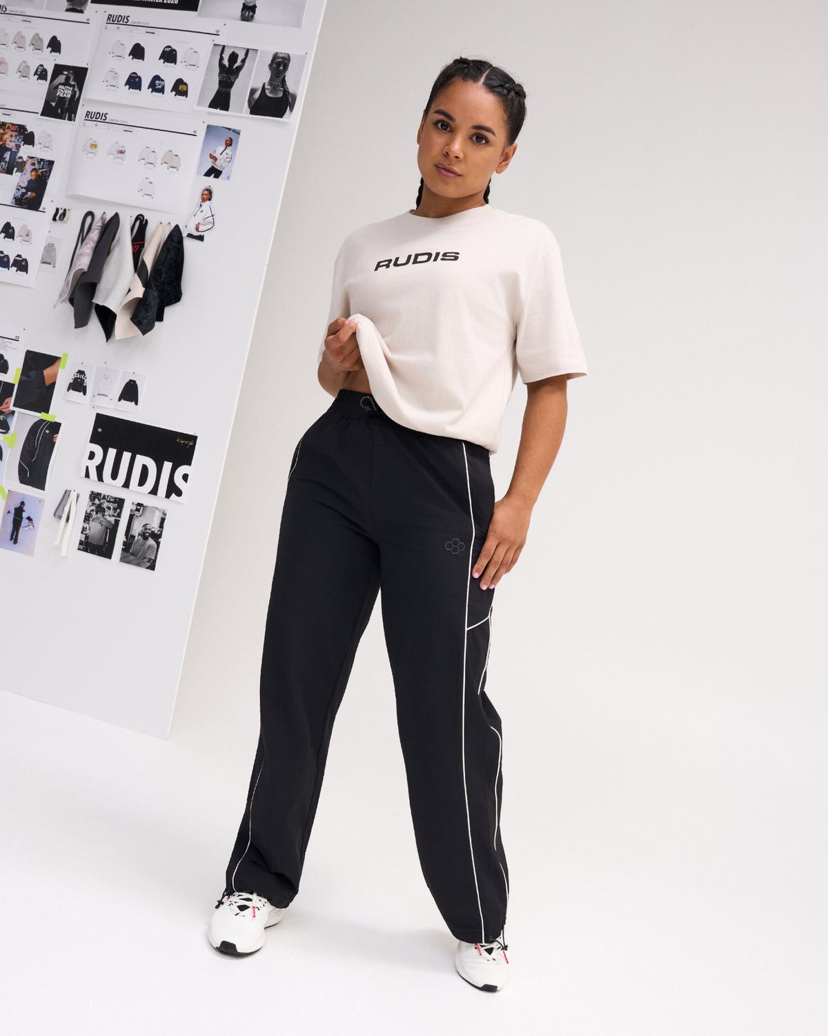 Female model wearing a bone RUDIS Relaxed Cotton T-Shirt with black RUDIS chest logo, paired with black athletic pants featuring white piping.