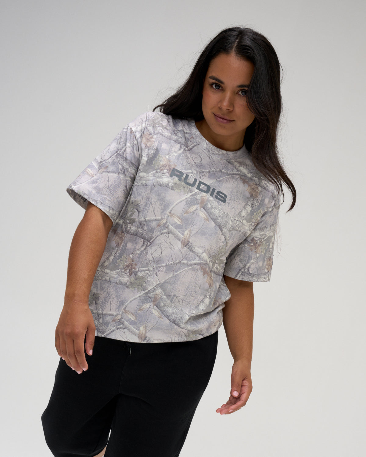Female model wearing the RUDIS Relaxed Cotton T-Shirt in light gray tree camo with a gray RUDIS chest logo and black athletic pants.