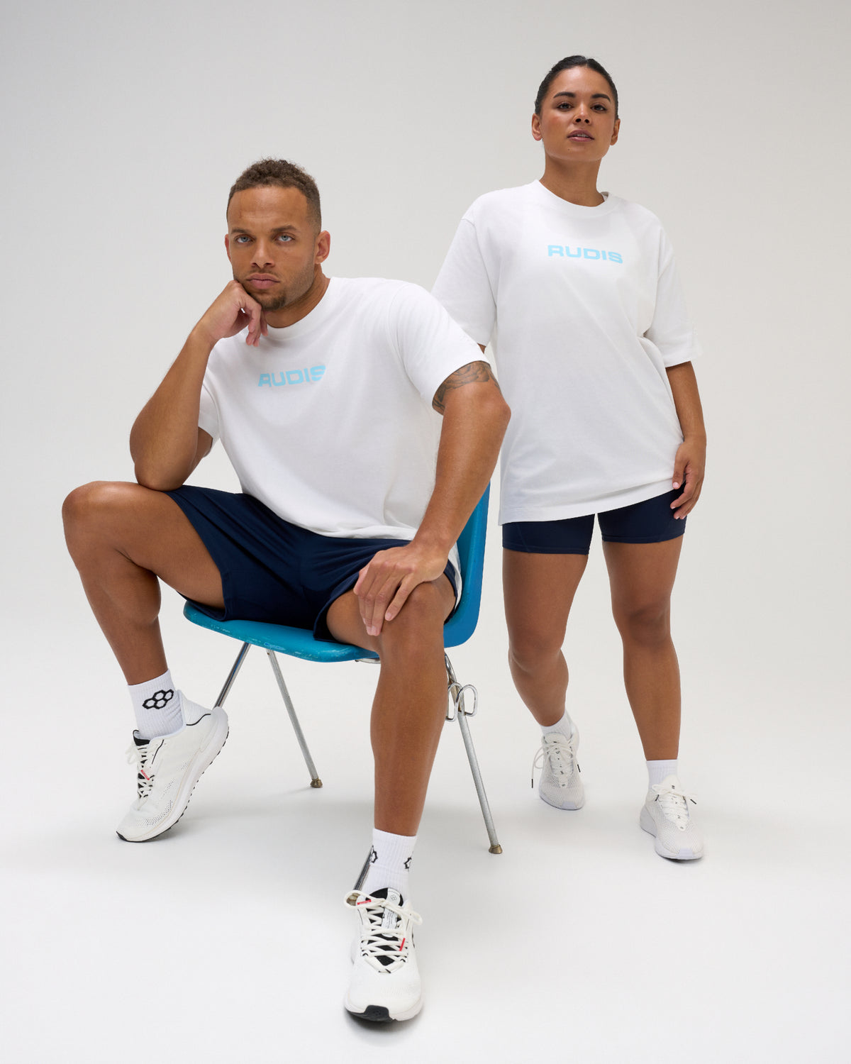 Male and female models wearing white RUDIS Relaxed Cotton T-Shirts with light blue RUDIS chest logos, paired with navy athletic bottoms.