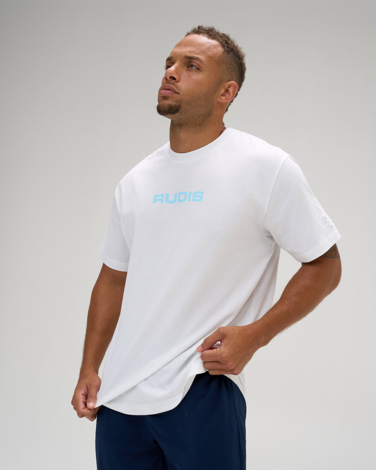 Male model wearing a white RUDIS Relaxed Cotton T-Shirt with a light blue RUDIS chest logo, paired with navy athletic shorts.