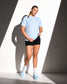 A woman modeling a light blue athletic t-shirt with white text on the chest, paired with black athletic shorts, white socks, and light blue sneakers, standing in dramatic diagonal sunlight against a white wall.