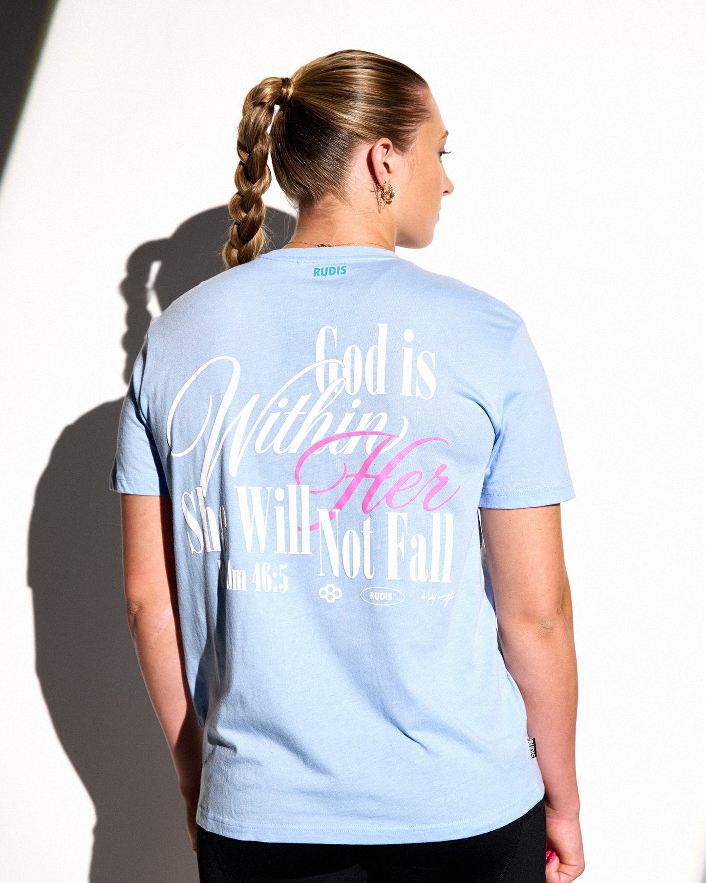 A woman shown from the back wearing a light blue graphic t-shirt with white and pink text that reads 'God is Within Her She Will Not Fall' and 'In 18:5'. The brand name 'BOBBI' is visible on the inner neck label.