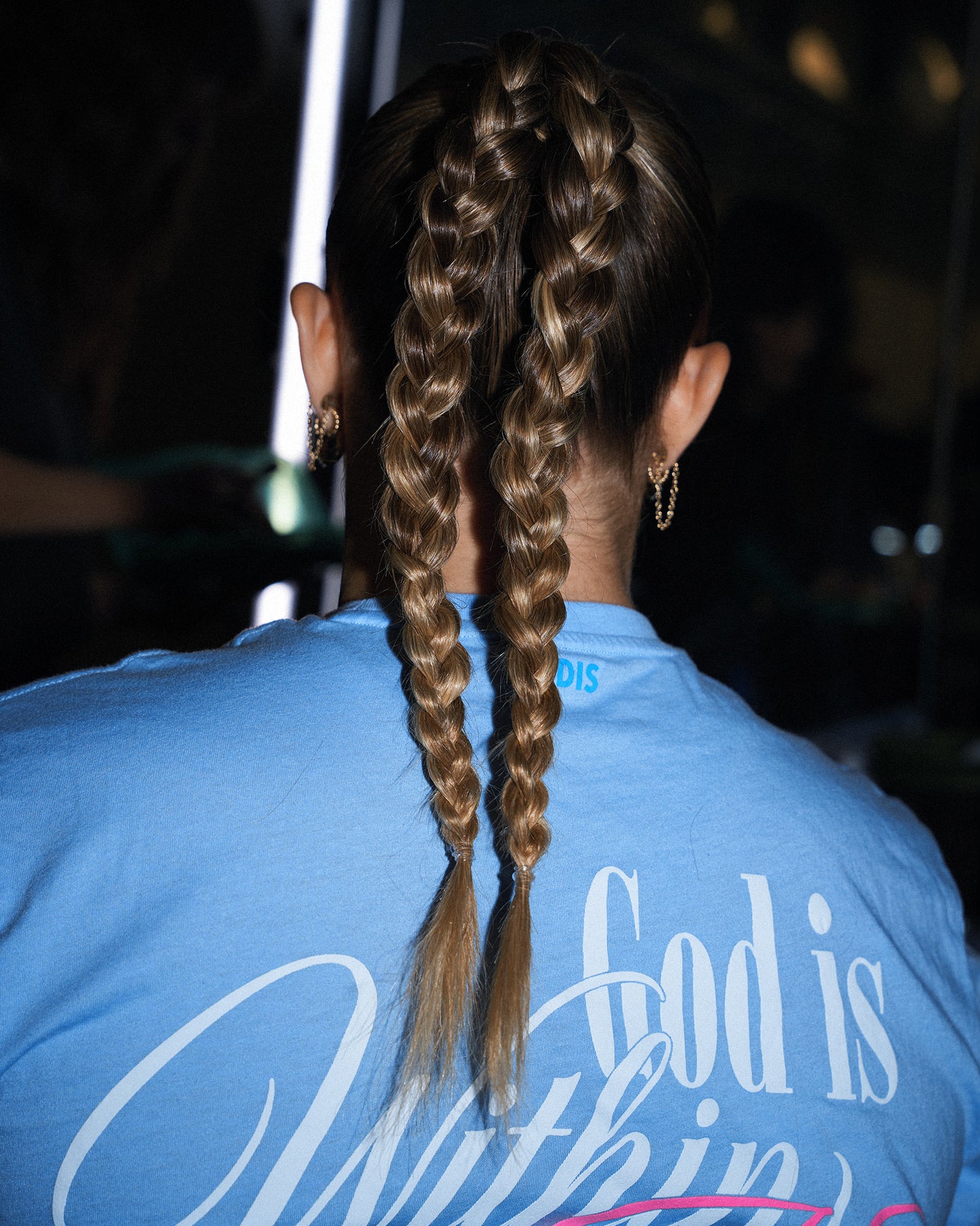 Back view of a person with long light brown hair styled in two braided ponytails, wearing a light blue t-shirt with 'God is Within' printed on the back.