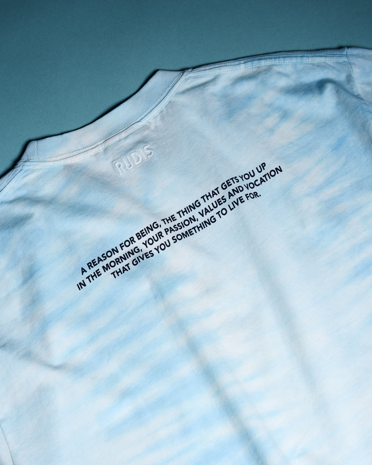 Close-up of the RUDIS Samurai Ikigai Classic Cotton T-Shirt in blue tie-dye, highlighting embroidered RUDIS branding and an inspirational message about purpose, passion, values, and vocation.