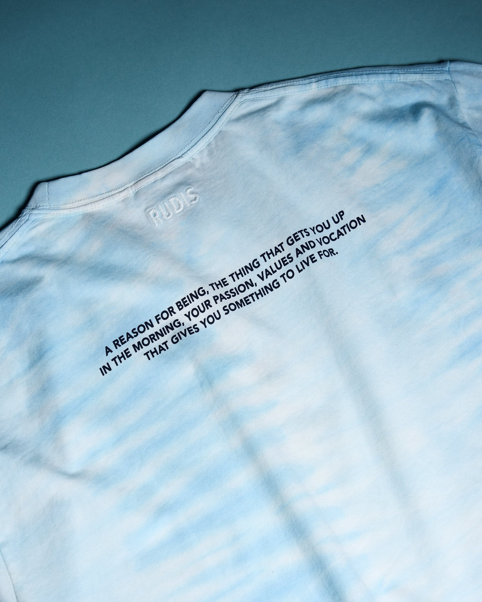 Close-up of the RUDIS Samurai Ikigai Classic Cotton T-Shirt in blue tie-dye, highlighting embroidered RUDIS branding and an inspirational message about purpose, passion, values, and vocation.