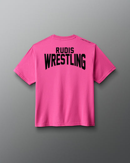 A bright pink short-sleeve t-shirt shown from the back, featuring the words 'RUDIS WRESTLING' printed in bold black capital letters across the upper back, set against a dark gray gradient background.