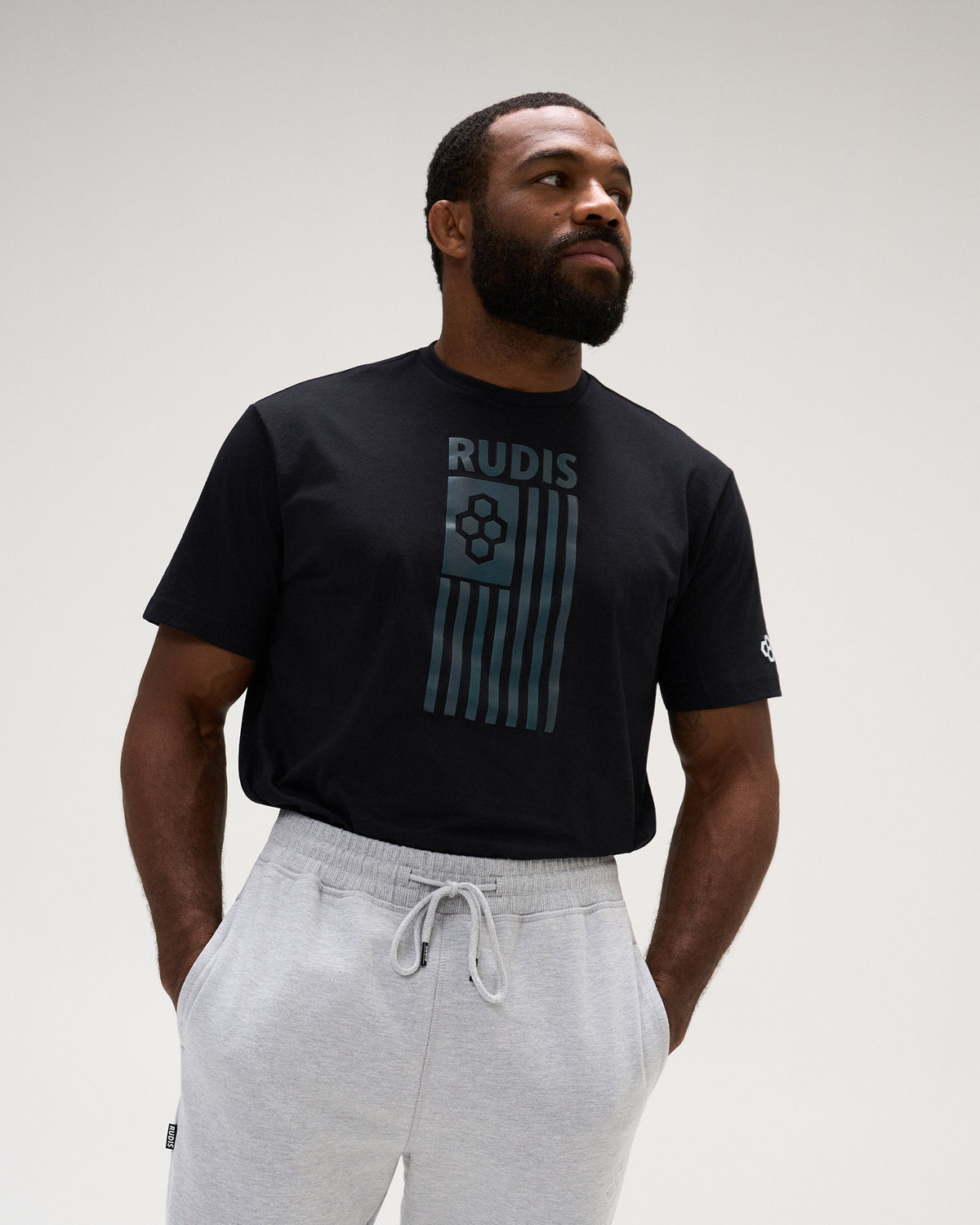 A man wearing a black short-sleeve t-shirt with a dark teal graphic on the front that reads 'RUDIS' above vertical stripes, paired with light gray sweatpants, against a plain light gray studio background.