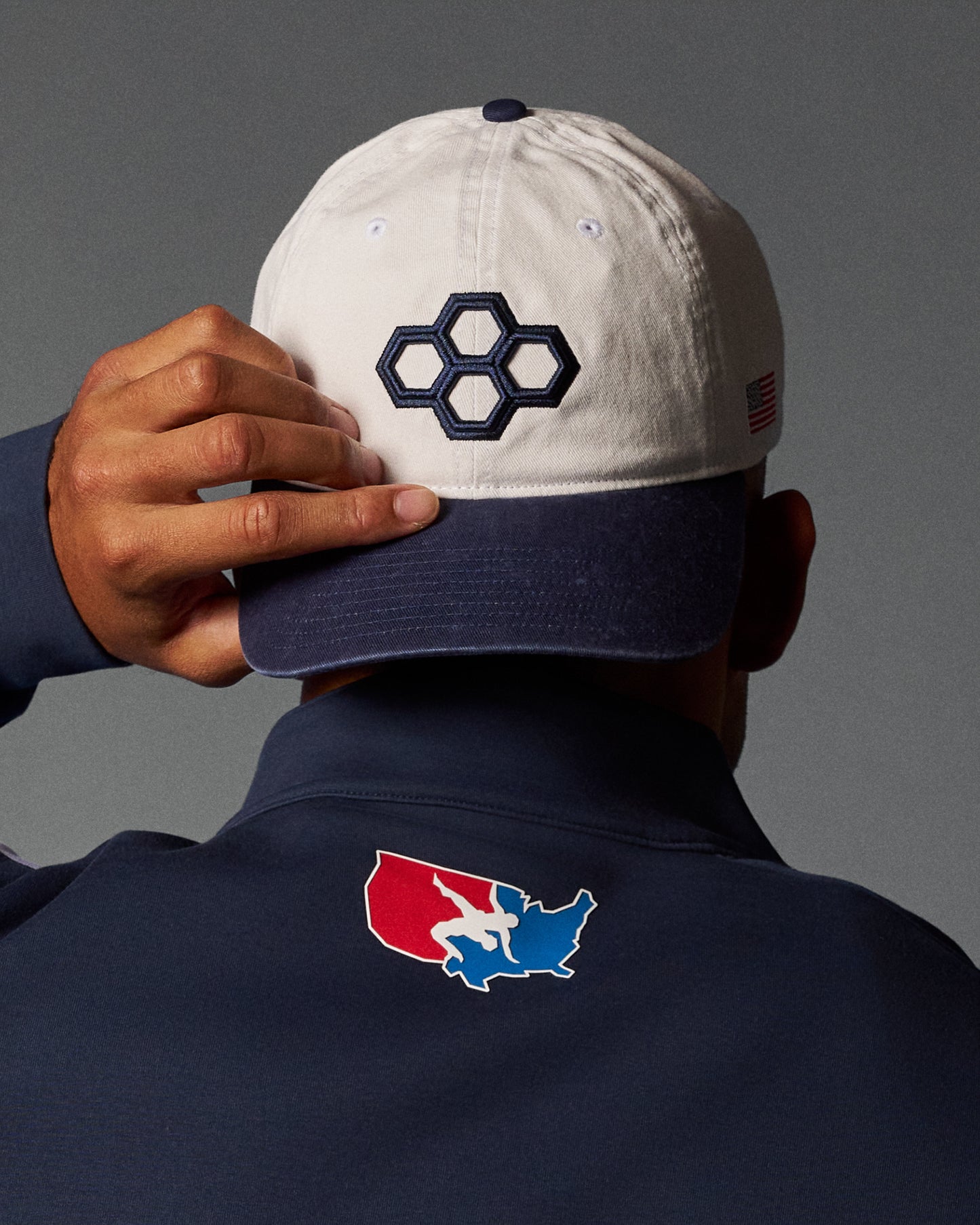 Person wearing a white cap with a hexagonal logo and navy jacket with a colorful logo on a gray background