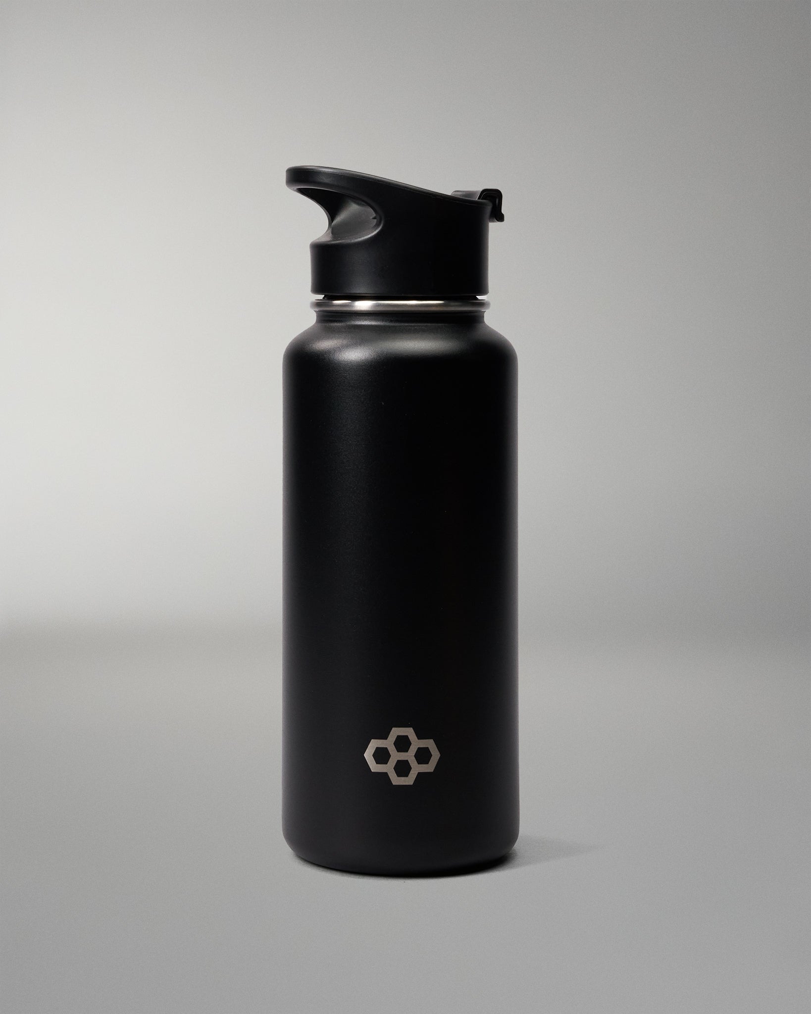 A matte black insulated water bottle with a matching black flip-top lid, featuring a small silver geometric logo near the bottom, set against a neutral gray studio background.
