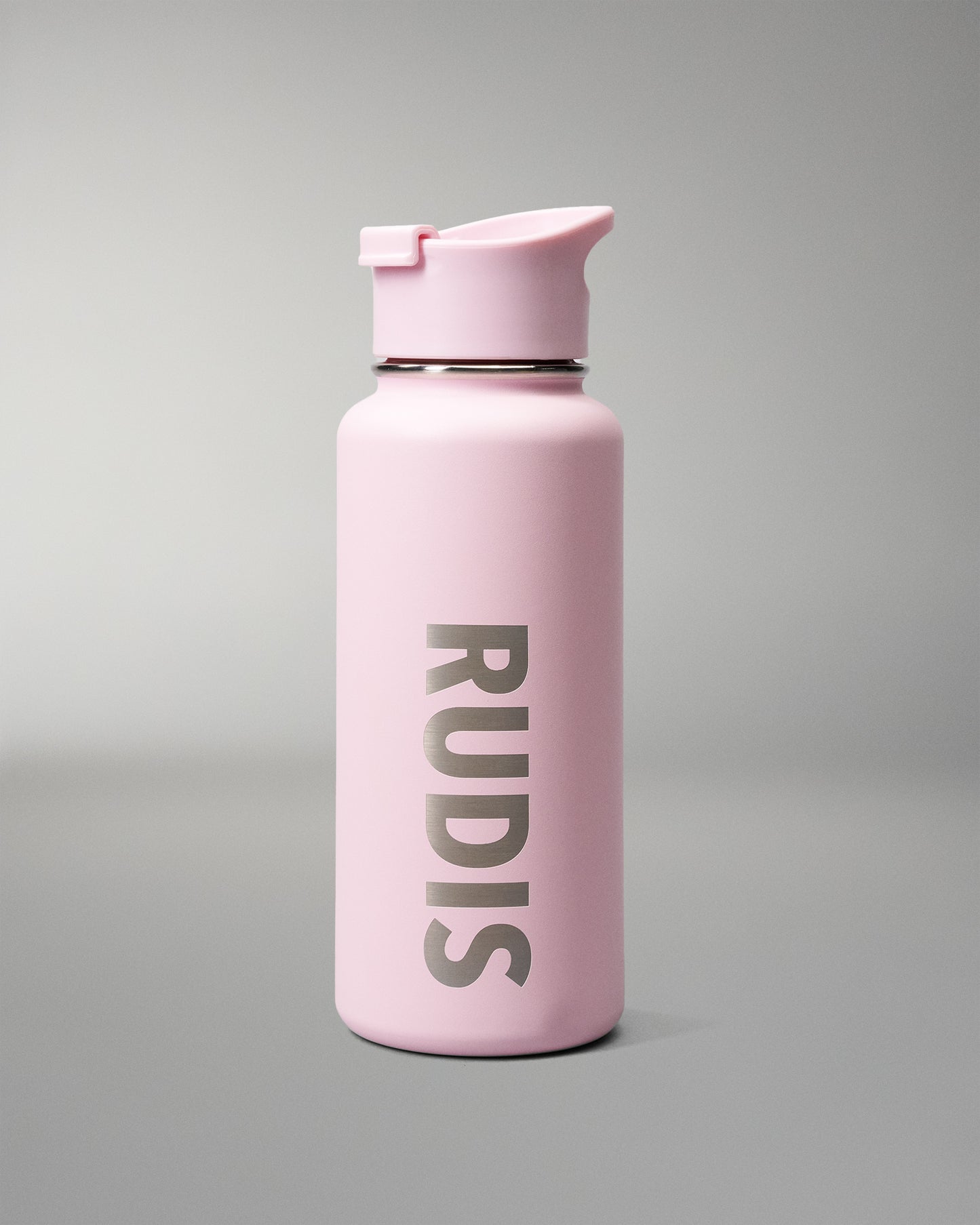 A matte light pink insulated water bottle with a matching flip-top lid, featuring the brand name 'RUDIS' printed vertically in dark gray on the front, set against a neutral gray studio background.