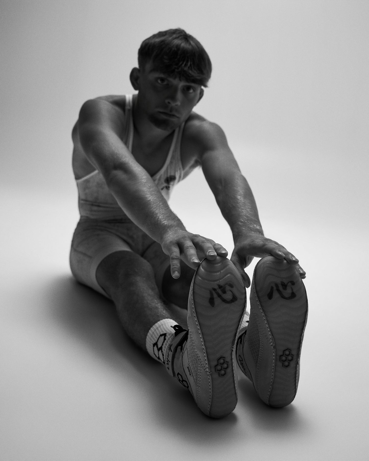 A black and white editorial photograph of a man in Wrestling singlet stretching, with his hands reaching toward his feet. He is wearing a wrestling singlet, socks, and wrestling shoes with a distinctive RUDIS logo on the soles.