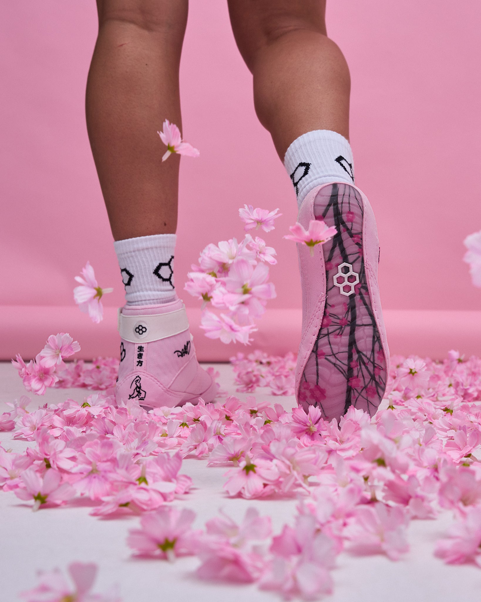 Rear view of an athlete wearing pink RUDIS wrestling shoes with a cherry blossom-inspired translucent outsole, white RUDIS socks, and pink blossoms scattered across the floor against a soft pink backdrop.