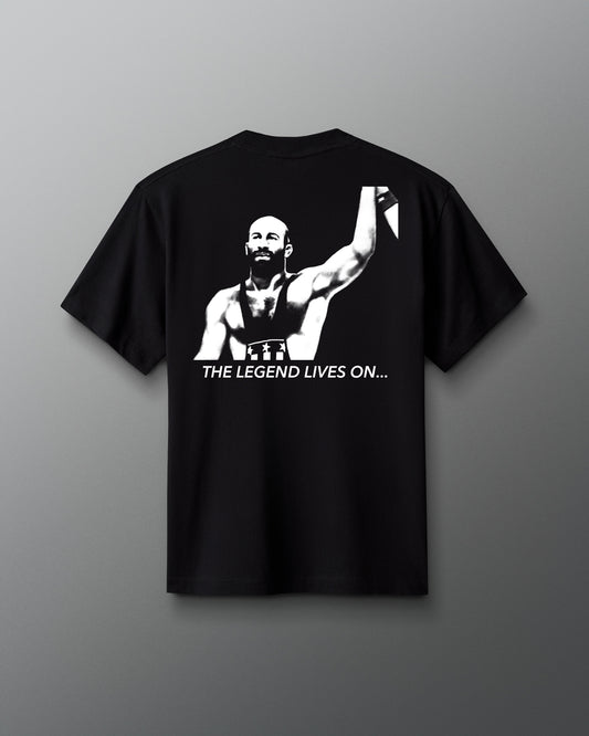Back view of a black t-shirt featuring a bold, high-contrast graphic of a victorious wrestler Dave Schultz raising his arm. Below the image, the phrase “THE LEGEND LIVES ON…” is printed in white capital letters.