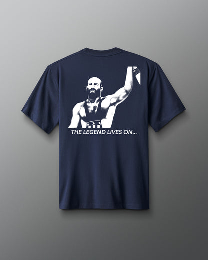 Back view of a navy t-shirt featuring a bold, high-contrast graphic of a victorious wrestler raising his arm. Below the image, the phrase “THE LEGEND LIVES ON…” is printed in white capital letters.