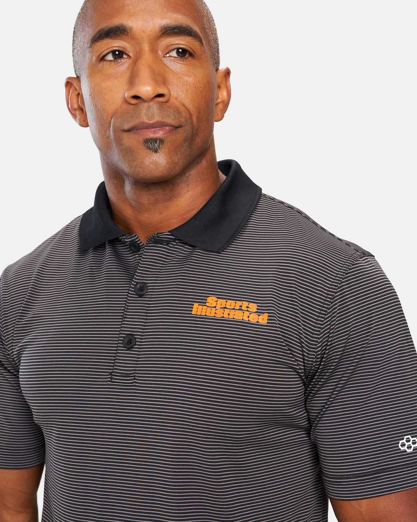 A man wearing a striped black polo shirt with an orange Sports Illustrated logo displaying a confident expression against a neutral background