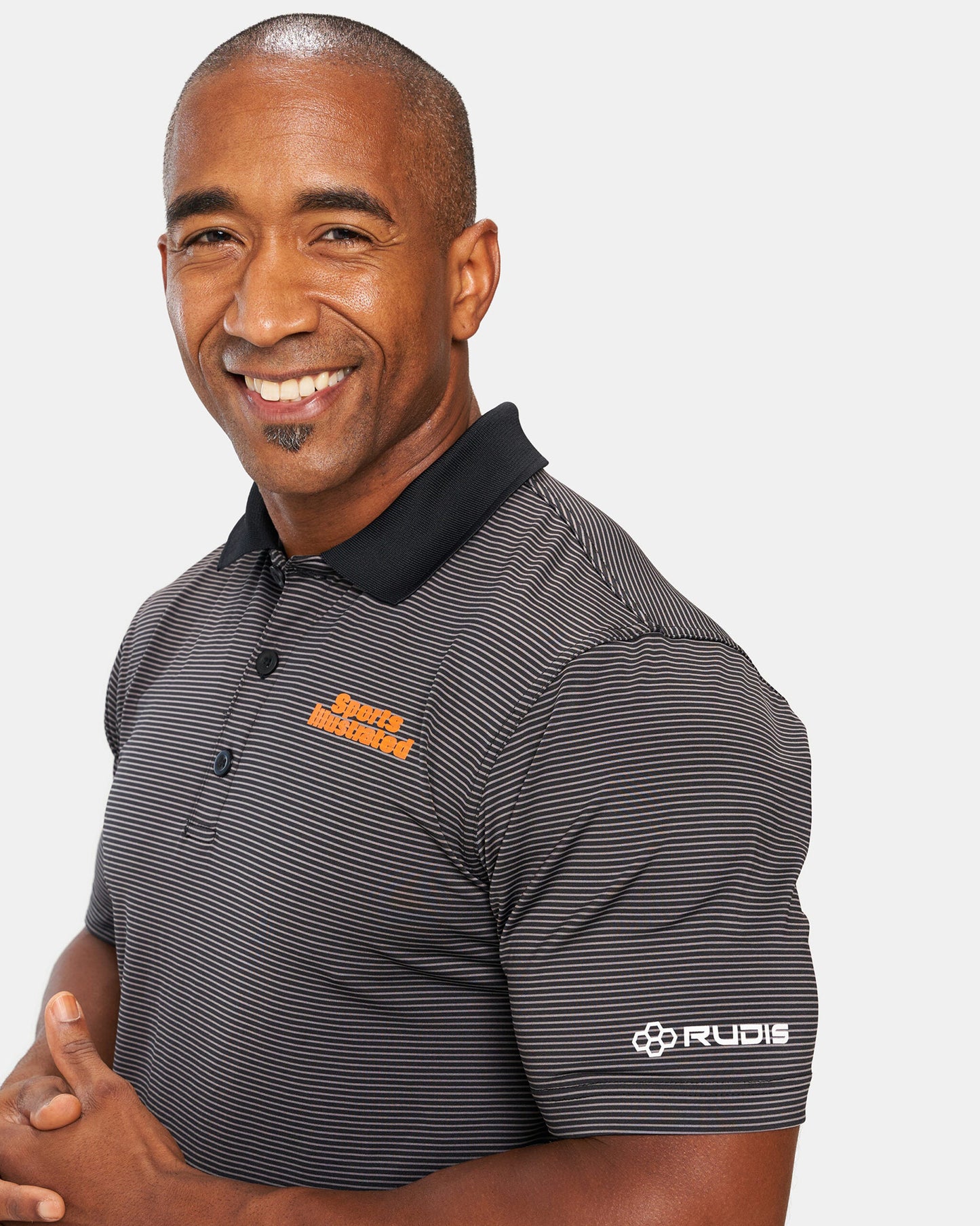 A portrait of a smiling man wearing a black and gray striped polo shirt with a Sports Illustrated logo and the Rudis brand name on the sleeve