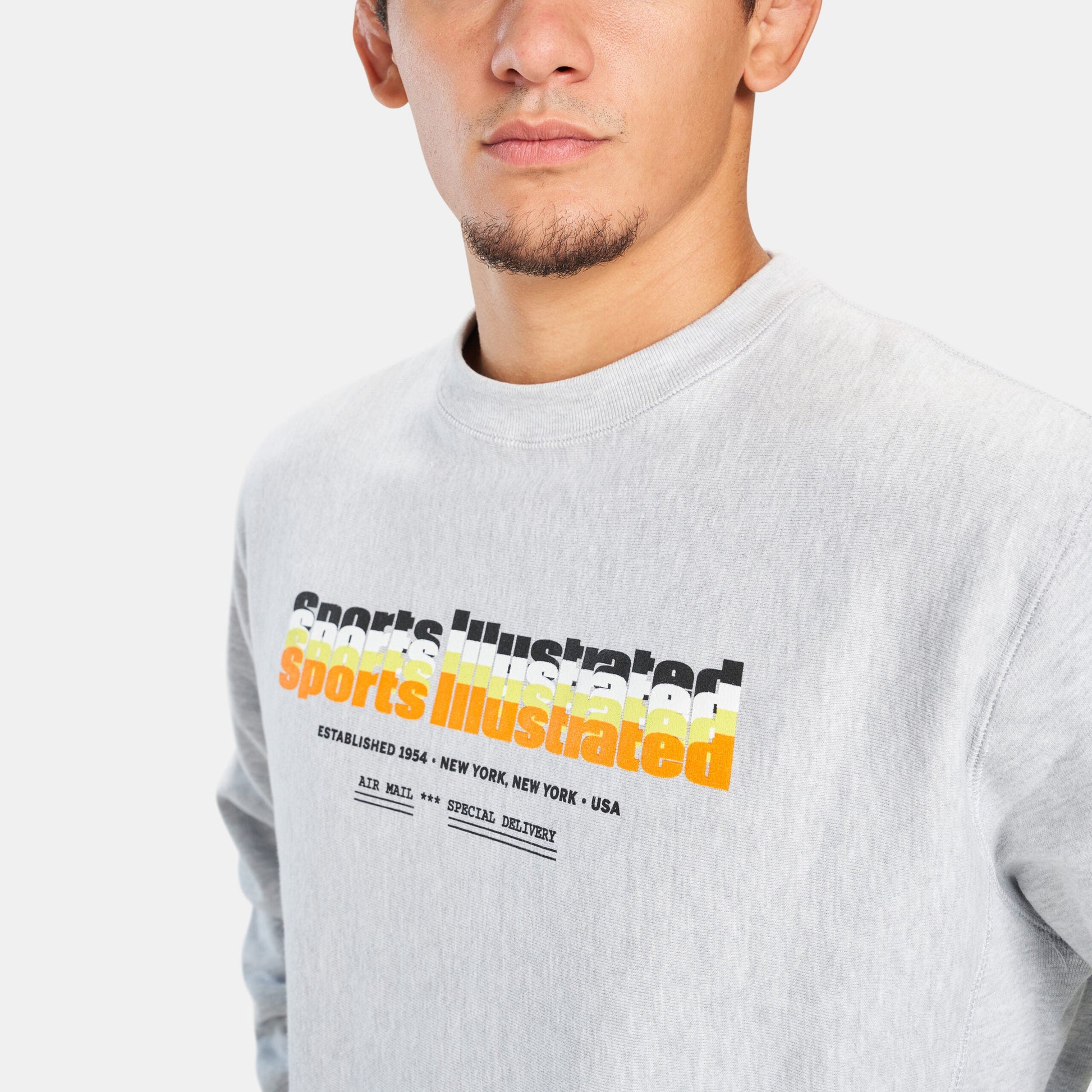 A close-up portrait of a young man wearing a light gray sweatshirt featuring a colorful graphic print of Sports Illustrated
